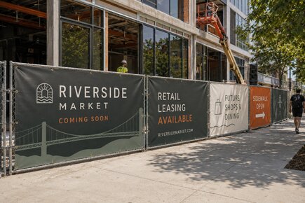Construction fence banners around a commercial development advertising riverside market retail leasing future shops and an open sidewalk