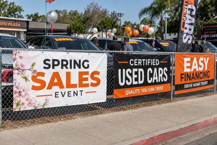 Fence banners advertising a car dealership spring sales event certified used cars and financing along a chain link perimeter