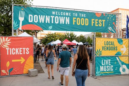 Large event fence banners marking the entrance to a downtown food festival with ticket vendor and live music information