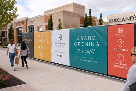 Commercial fence banners outside a shopping center promoting new stores, a grand opening, parking and current entrance directions