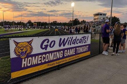 Custom school vinyl banner promoting a Wildcats homecoming football game mounted on a chain link stadium fence